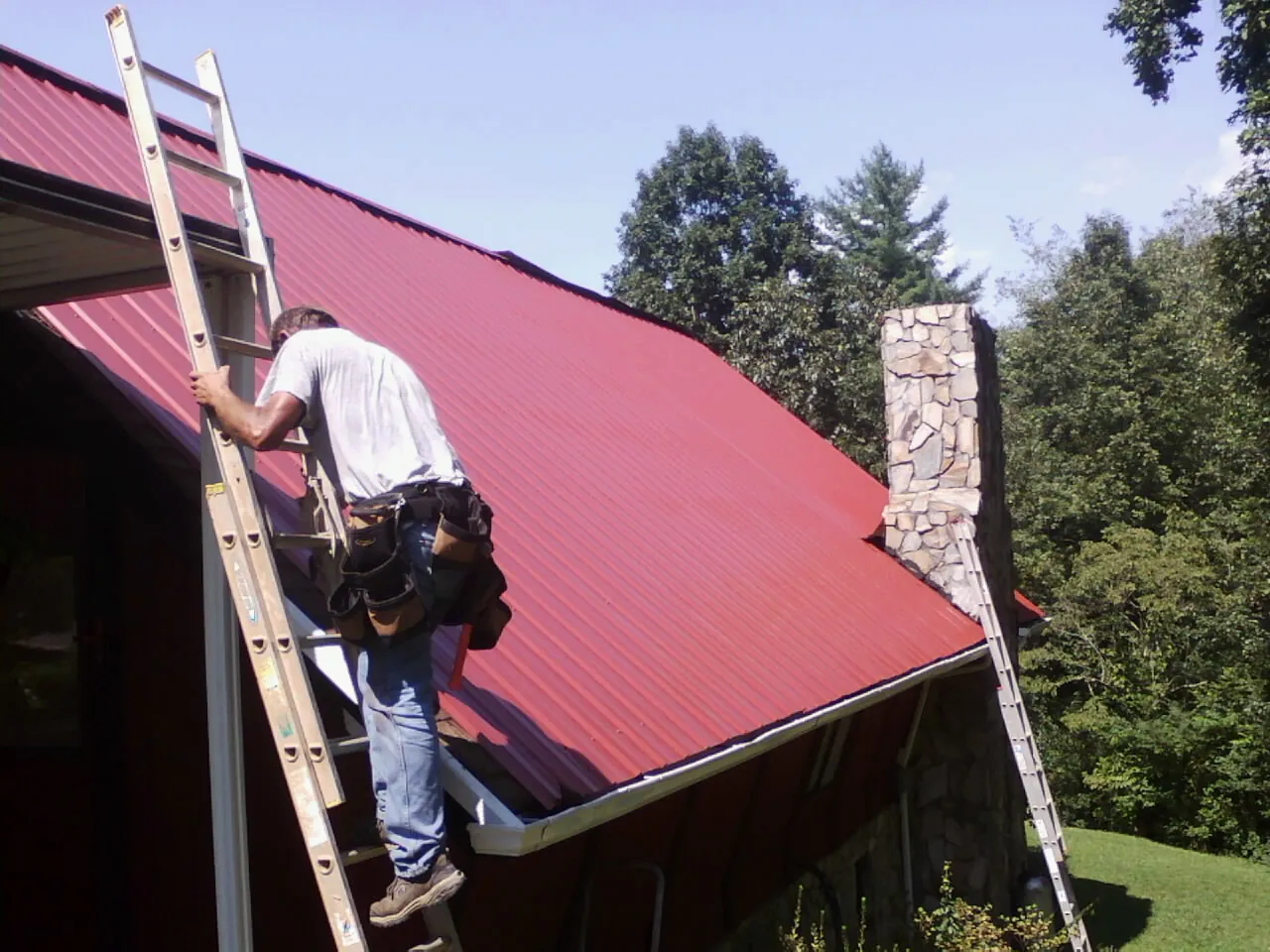 Roofer on a ladder beside a red metal roof and stone chimney — Soffit & Fascia Repair by Roofing Brothers in Sedalia